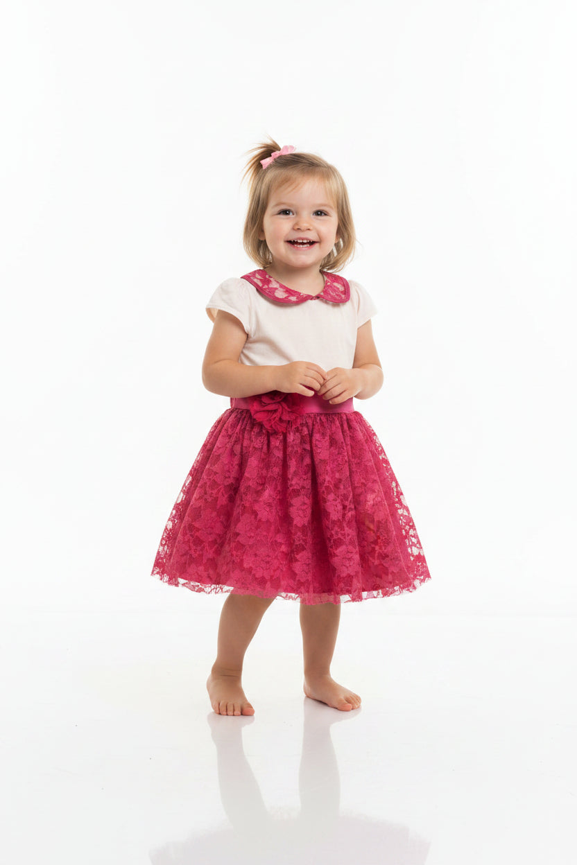 A girls' dress hanging on a white hanger against a plain light gray wall. The dress has a cream-colored top with a pink floral lace Peter Pan collar, short cap sleeves, a pink satin waistband, and a full pink floral lace skirt with a large fabric flower on the left side.