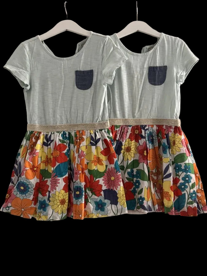 Two matching girls' dresses hanging on white hangers against a plain light gray wall. Each dress has a pale mint green short-sleeve top with a dark blue pocket and a colorful floral skirt with orange, yellow, red, blue, and green flowers.
