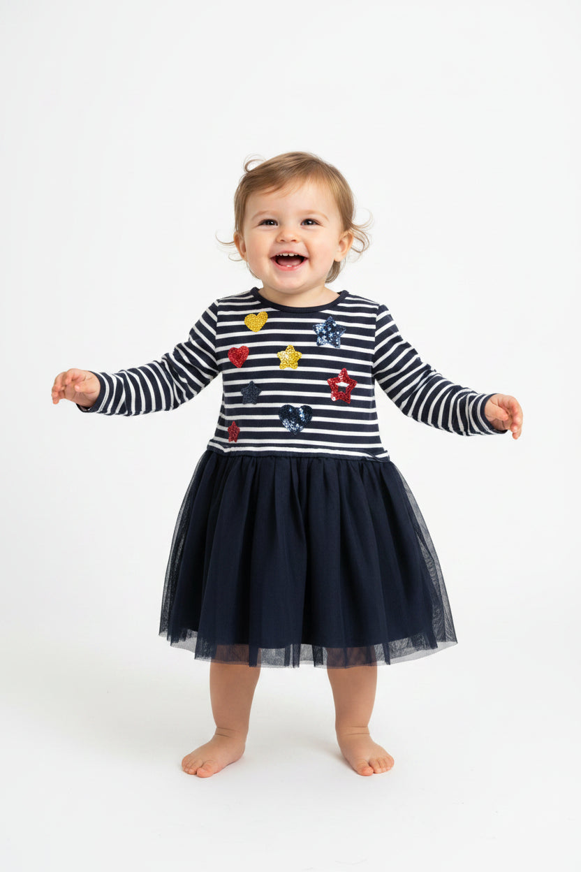A baby girl's long-sleeve dress hanging on a white hanger against a plain light gray wall. The top half features navy and white horizontal stripes with colorful sequined heart and star patches, and the bottom half is a dark navy blue tulle skirt.