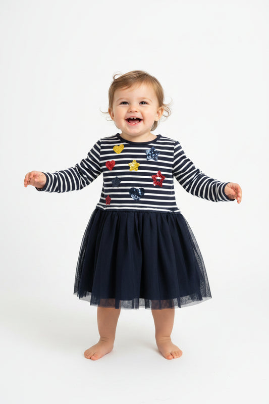 A baby girl's long-sleeve dress hanging on a white hanger against a plain light gray wall. The top half features navy and white horizontal stripes with colorful sequined heart and star patches, and the bottom half is a dark navy blue tulle skirt.