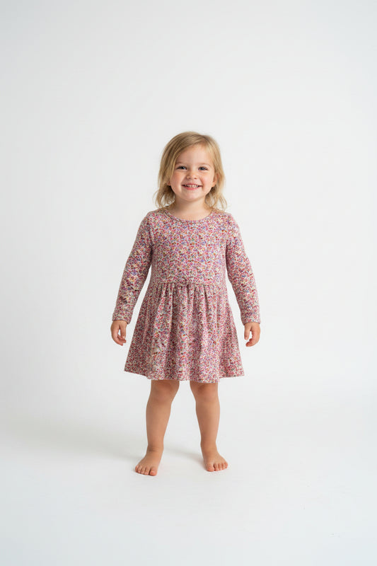 A long-sleeve children's dress with a dense small floral print in pink, red, and blue on a light base, hanging on a white plastic hanger against a plain light gray wall.