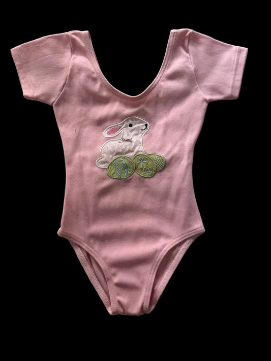 A pink short-sleeve baby bodysuit featuring an embroidered white bunny and green Easter eggs on the chest, laid flat on a textured gray fabric background.