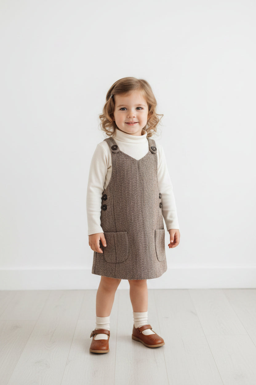 A sleeveless brown tweed pinafore dress with a windowpane check pattern, featuring shoulder straps with decorative buttons, side buttons, and two front patch pockets, hanging on a white hanger against a light gray wall.